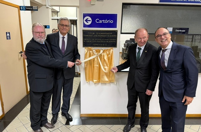 Foto horizontal: autoridades de preto em cartório branco, ambiente institucional, inaugurando placa com faixa dourada
