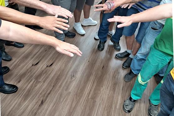 Foto horizontal: pessoas estendem as mãos em dinâmica coletiva sobre piso amadeirado em ambiente interno.