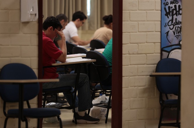 Foto horizontal: candidatos realizam prova em sala de aula vista por corredor com paredes claras.