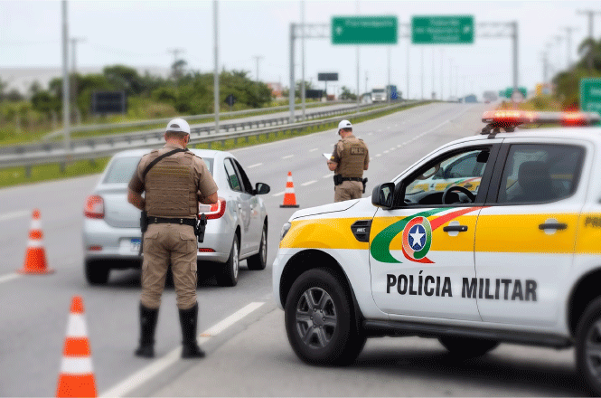 Foto horizontal: policiais militares fiscalizam veículos em rodovia com cones e viatura ao fundo.