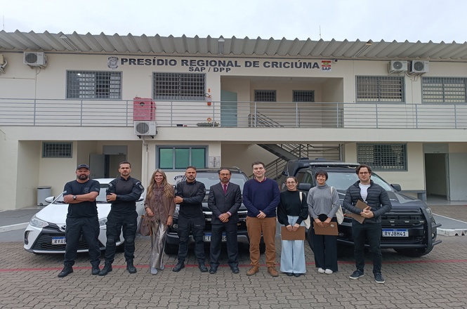 Foto horizontal: grupo posa diante do Presídio Regional de Criciúma ao lado de viaturas e agentes.