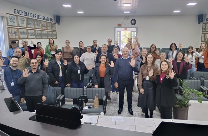 Foto horizontal: grupo de pessoas acena em plenário institucional sob iluminação branca e tons cinza.