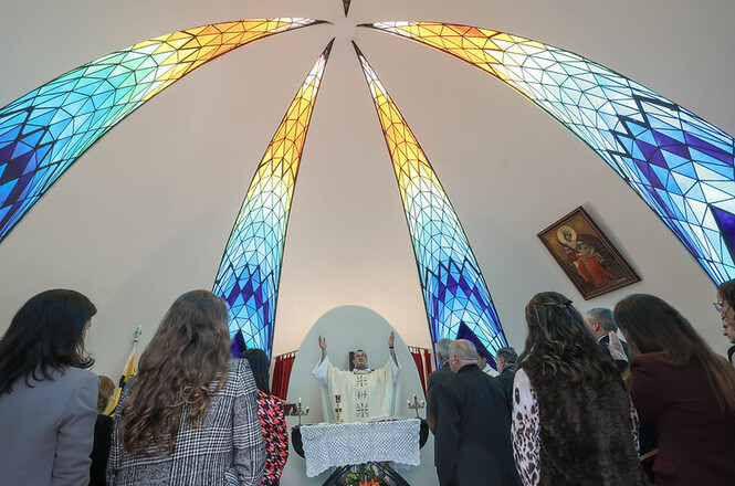 Foto horizontal: fiéis acompanham missa sob vitrais coloridos em capela de tons azul e dourado.