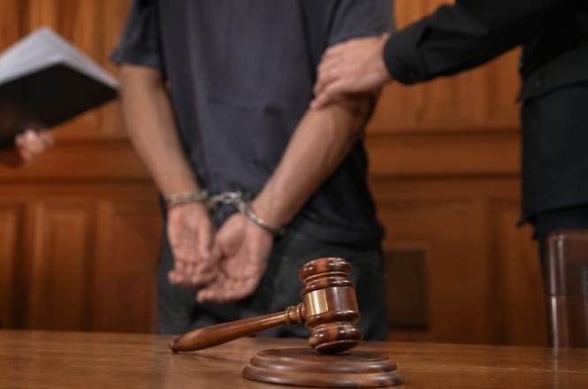Foto horizontal: pessoa de preto algemada em tribunal marrom, ambiente judicial, sendo contida junto a juiz e martelo