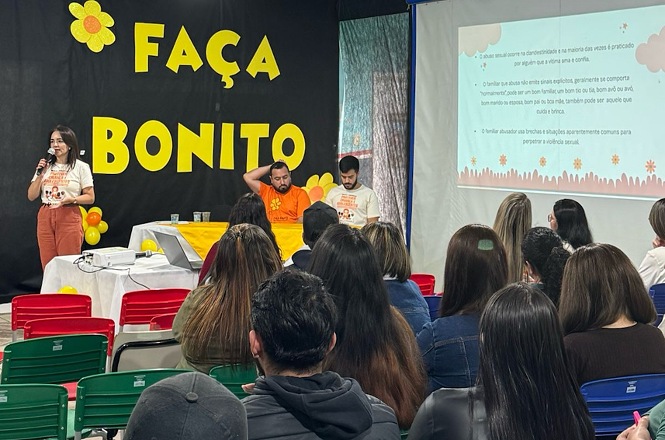 Foto horizontal: pessoas em tons amarelo e preto, sala com painel colorido, educativo, palestrando para público sentado