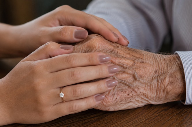 Foto horizontal: mãos jovens seguram mãos idosas em gesto de cuidado e afeto, detalhe em anel, tons suaves.