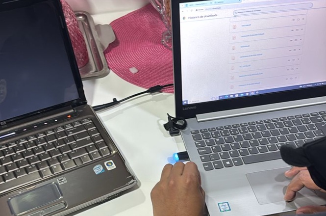 Foto horizontal: mãos utilizam notebooks conectados, navegação em sistema digital, ambiente doméstico, tons claros.