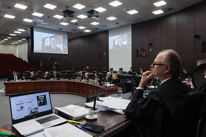 Foto horizontal: desembargador Rubens Schulz acompanha sessão com videoconferência ao fundo, magistrados em plenário, ambiente institucional.