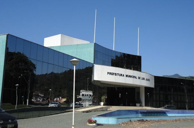Foto horizontal: fachada da Prefeitura Municipal de Luis Alves, prédio moderno com vidro espelhado, área externa e céu azul.