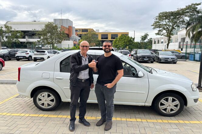 Foto horizontal: dois homens posam ao lado de veículo estacionado, cumprimento em destaque, ambiente externo, tons neutros.