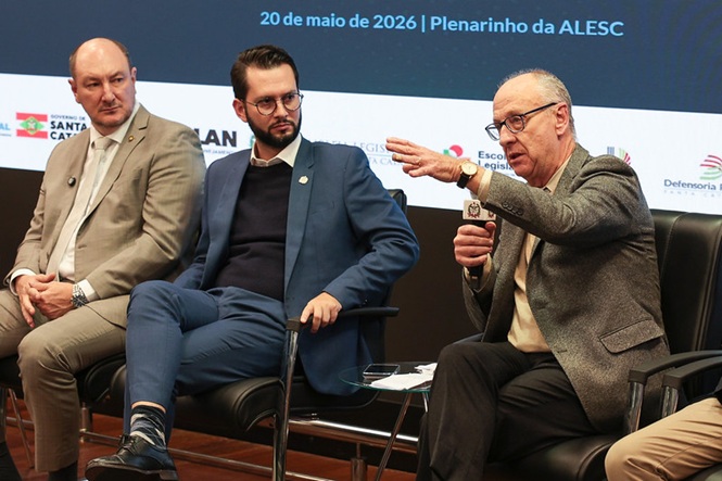 Foto horizontal: participantes debatem em painel na ALESC, um ao microfone com gesto em destaque, outros atentos, ambiente institucional.