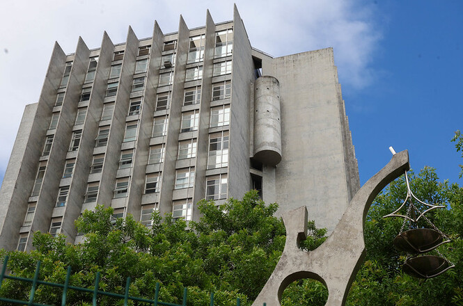 Foto horizontal: edifício do TJSC em arquitetura moderna, estrutura em concreto com linhas verticais, vegetação ao redor, céu parcialmente nublado.