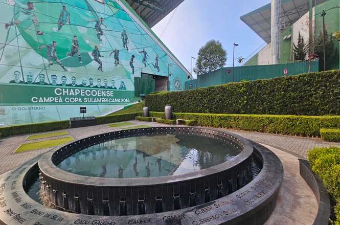 Foto horizontal: memorial da Chapecoense com fonte circular em primeiro plano e mural ao fundo, homenagem às vítimas, ambiente externo, tons verdes.