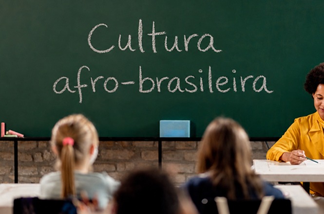 Imagem horizontal: quadro com a frase “Cultura afro-brasileira” ao fundo, alunos de costas e professora em sala de aula, ambiente educacional.