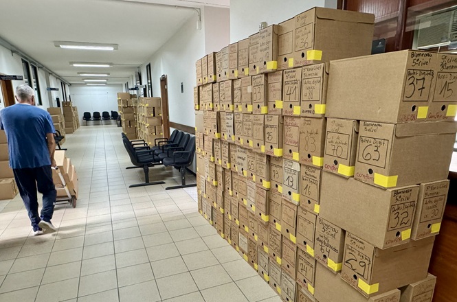 Foto horizontal: corredor com caixas organizadas e etiquetadas empilhadas, pessoa caminha ao fundo, ambiente interno, tons neutros.