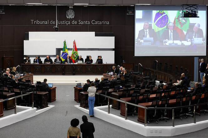 Foto horizontal: sessão de julgamento no plenário do TJSC, magistrados à mesa, bandeiras ao centro e telão ao fundo, ambiente institucional.