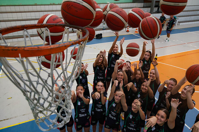 Foto horizontal: jovens atletas lançam bolas de basquete em quadra, cesta em primeiro plano, atividade esportiva coletiva, tons vibrantes.