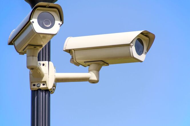 Foto horizontal: câmeras de vigilância instaladas em poste, monitoramento em espaço externo, céu azul ao fundo, tons claros.