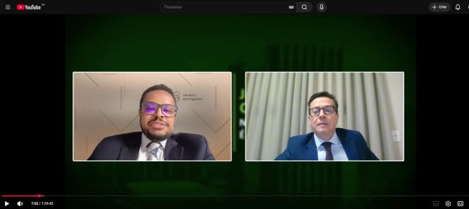 Descrição de acessibilidade: captura de tela de uma transmissão virtual no YouTube com dois participantes lado a lado. À esquerda, o palestrante Leonardo Braga Moura aparece de terno escuro, camisa branca e gravata, usando óculos de armação escura e fones de ouvido. Ao fundo, há uma parede clara com detalhes geométricos. À direita, o desembargador Edir Beck veste terno azul-marinho, camisa clara e gravata vinho, diante de uma cortina bege. A interface do YouTube aparece em modo escuro, com a barra de reprodução visível na parte inferior da imagem.