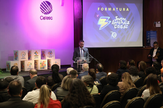 Foto horizontal: cerimônia de formatura do programa Jovem Aprendiz Celesc, orador ao púlpito e público presente, iluminação cênica, ambiente institucional.