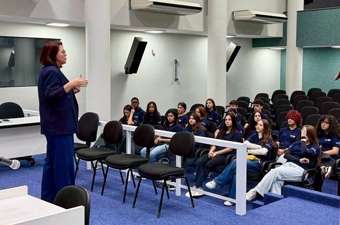 Foto horizontal: mulher conduz apresentação para grupo de jovens em sala institucional, plateia atenta, ambiente educacional, tons neutros.