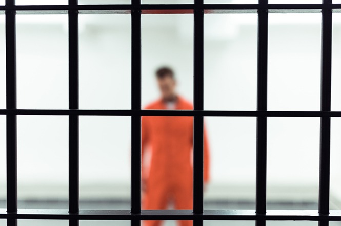 Foto horizontal: homem desfocado com uniforme laranja visto atrás de grades, referência ao sistema prisional, ambiente interno, tons frios.