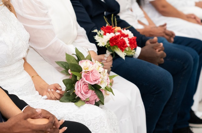 Foto horizontal: pessoas sentadas lado a lado em cerimônia de casamento, noivas com buquês em destaque, clima afetivo, tons claros.