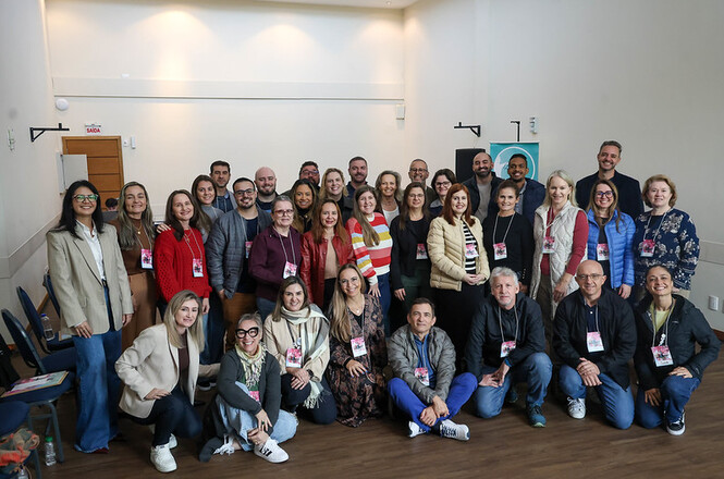 Foto horizontal: grupo de profissionais posa em ambiente interno, participantes com credenciais reunidos para atividade coletiva, clima colaborativo, tons neutros.