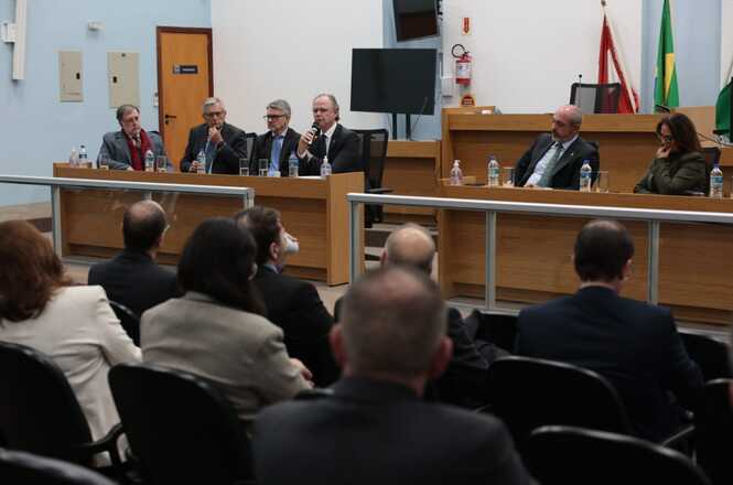 Foto horizontal: sessão institucional com autoridades em mesa, desembargador Rubens Schulz fala ao microfone, público acompanha ao fundo, ambiente formal.