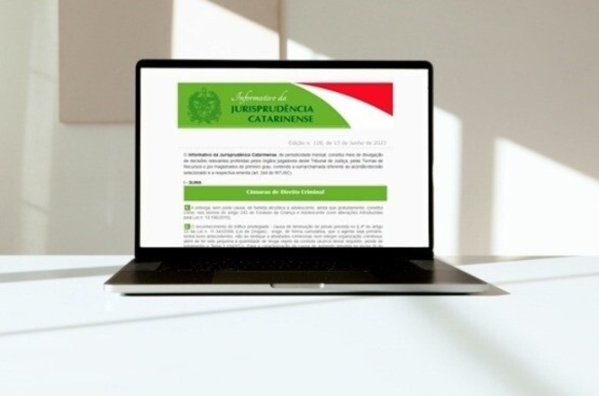 Foto horizontal: notebook exibe página de jurisprudência catarinense na tela, dispositivo sobre mesa em ambiente claro, foco em tecnologia jurídica, tons neutros.