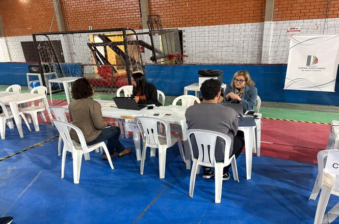 Foto horizontal: atendimento do projeto Justiça até Você em quadra esportiva, profissionais orientam cidadãos em mesas, ambiente comunitário, clima de acolhimento.