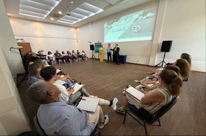 Foto horizontal: participantes em círculo acompanham apresentação com projeção ao fundo, facilitadores ao centro, ambiente amplo, clima de escuta e aprendizagem.
