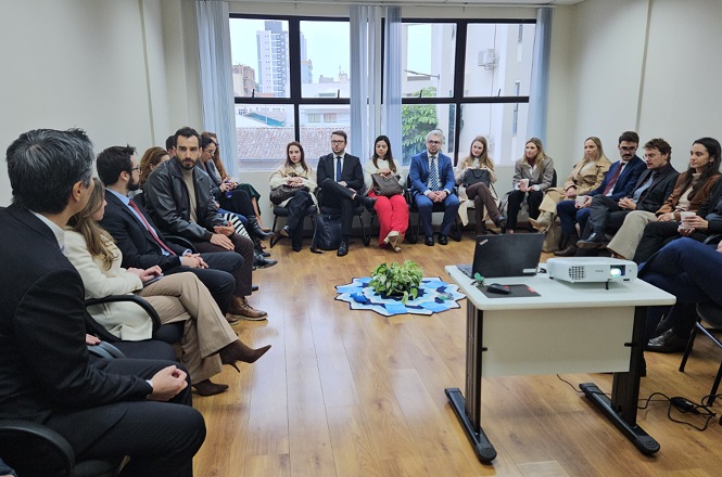 Foto horizontal: reunião institucional com participantes sentados em círculo, diálogo em andamento, ambiente interno com luz natural, clima de escuta e colaboração.