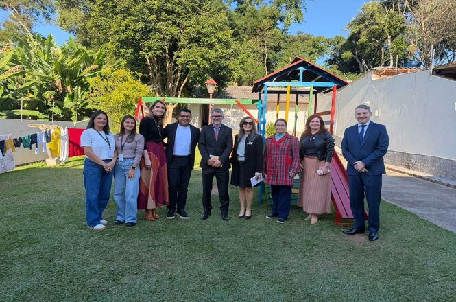 Foto horizontal: grupo de autoridades e equipe posa em área externa de casa de acolhimento, playground ao fundo, ambiente comunitário, clima institucional e acolhedor.