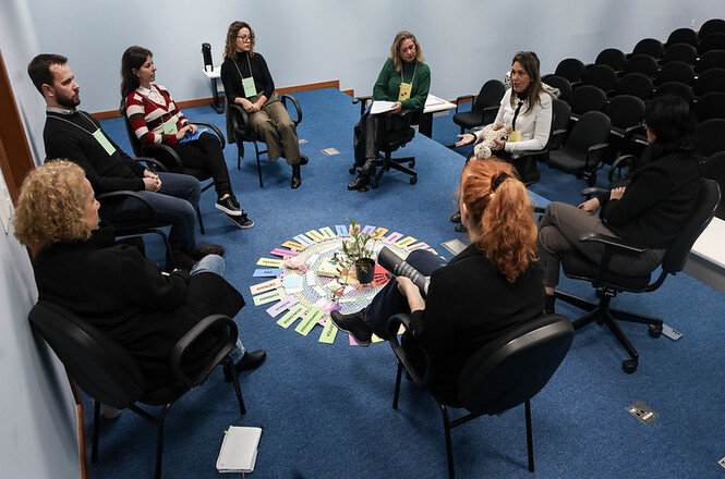 Foto horizontal: participantes sentados em círculo durante dinâmica de grupo, materiais dispostos no centro, ambiente interno, clima de escuta e interação.