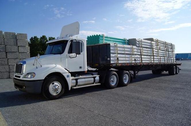 Foto horizontal: caminhão de carga estacionado com carroceria aberta transportando materiais, área industrial ao fundo, ambiente externo, tons neutros.