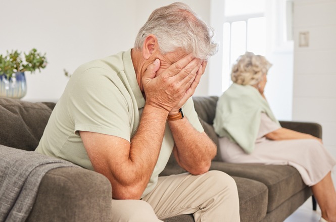 Foto horizontal: homem idoso cobre o rosto com as mãos em expressão de sofrimento, mulher ao fundo sentada no sofá, ambiente doméstico, clima de tensão e tristeza.