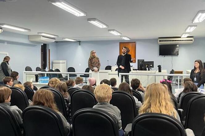 Foto horizontal: magistrados conversam com estudantes em plenário, atividade educativa em andamento, público jovem atento, ambiente institucional.