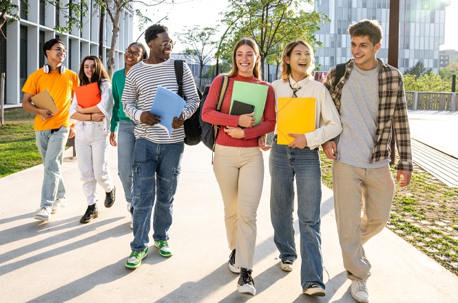 Foto horizontal: grupo de estudantes caminha ao ar livre com cadernos e mochilas, interação descontraída, ambiente universitário, clima de convivência e aprendizado.