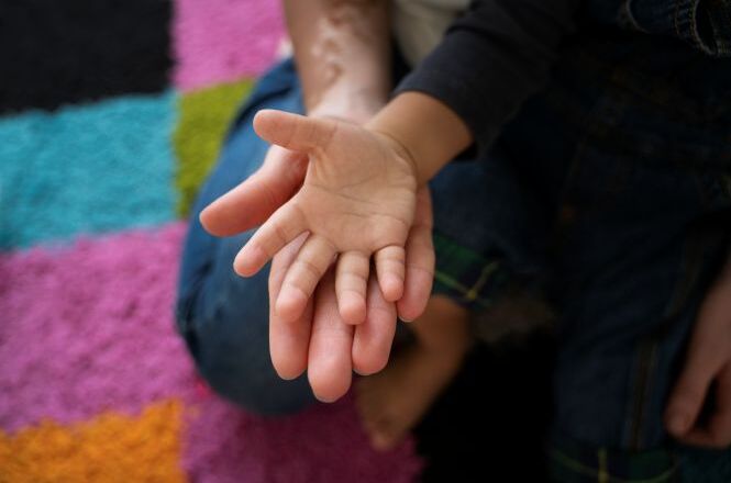 Foto horizontal: mão de criança apoiada sobre a mão de adulto, gesto de cuidado e proteção, ambiente acolhedor ao fundo, clima de afeto e desenvolvimento.