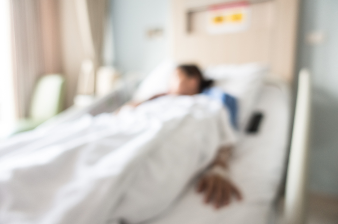 Foto horizontal: paciente deitado em leito hospitalar, imagem desfocada, ambiente clínico ao redor, clima de fragilidade e cuidado médico.