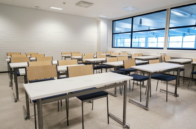 Foto horizontal: sala de aula vazia com mesas e cadeiras organizadas, ambiente iluminado por luz natural, espaço institucional, clima de silêncio e preparação.