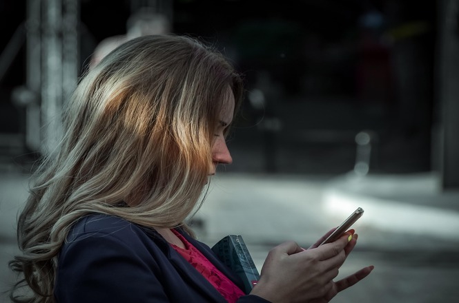Foto horizontal: mulher utiliza celular em ambiente externo, foco no rosto iluminado pela tela, fundo desfocado, clima de concentração e conexão digital.