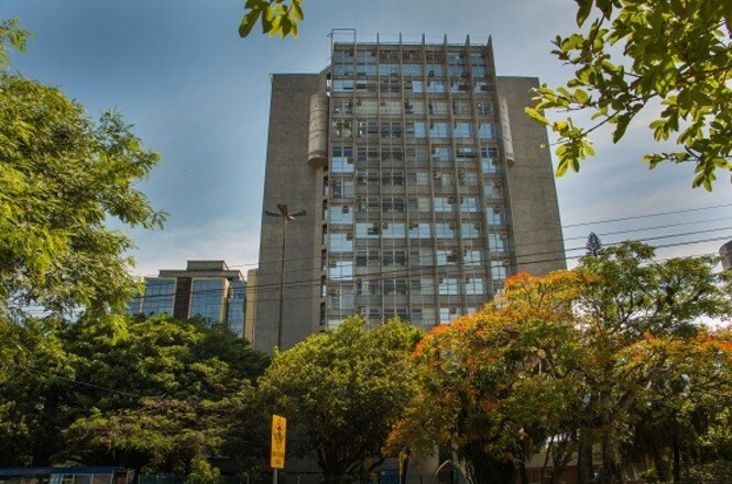 Foto horizontal: prédio cinza e vidro com árvores verdes e céu azul, área urbana, destacando sede do TJSC