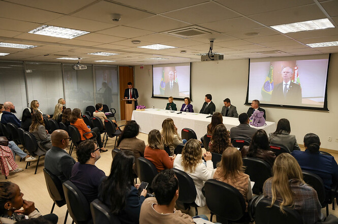Foto horizontal: público acompanha mesa de debate em evento institucional, autoridades à frente e projeção em telão ao fundo, ambiente formal, clima de reflexão e diálogo coletivo.