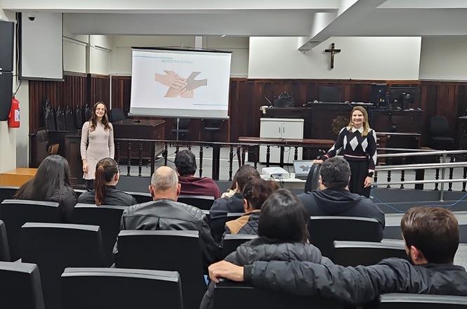 Foto horizontal: participantes acompanham atividade educativa em sala institucional, facilitadoras ao frente com apresentação projetada, público atento, ambiente formal, clima de aprendizagem e interação.