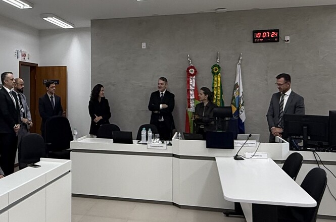 Foto horizontal: autoridades e servidores conversam durante inspeção em sala institucional, bandeiras ao fundo e ambiente formal, clima de alinhamento e cooperação.