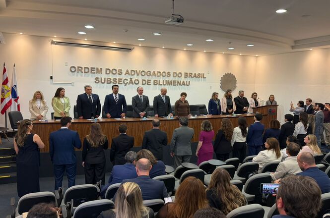 Foto horizontal: autoridades participam de solenidade na OAB Subseção de Blumenau, dispostas em mesa diretiva enquanto público acompanha em pé, ambiente institucional, clima de formalidade e respeito.