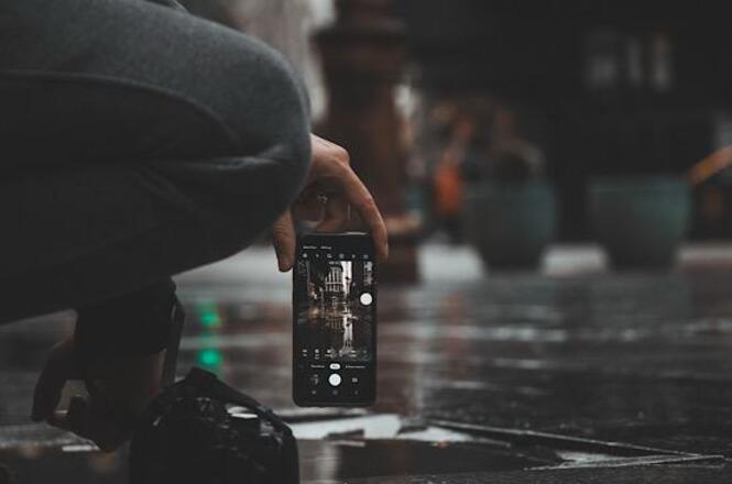 Foto horizontal: pessoa fotografa com celular em ambiente urbano molhado, reflexos no chão, clima de registro e cotidiano.