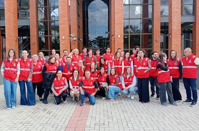 Foto horizontal: equipe com coletes vermelhos posa em frente a prédio institucional, registro coletivo, clima de organização e atuação conjunta.